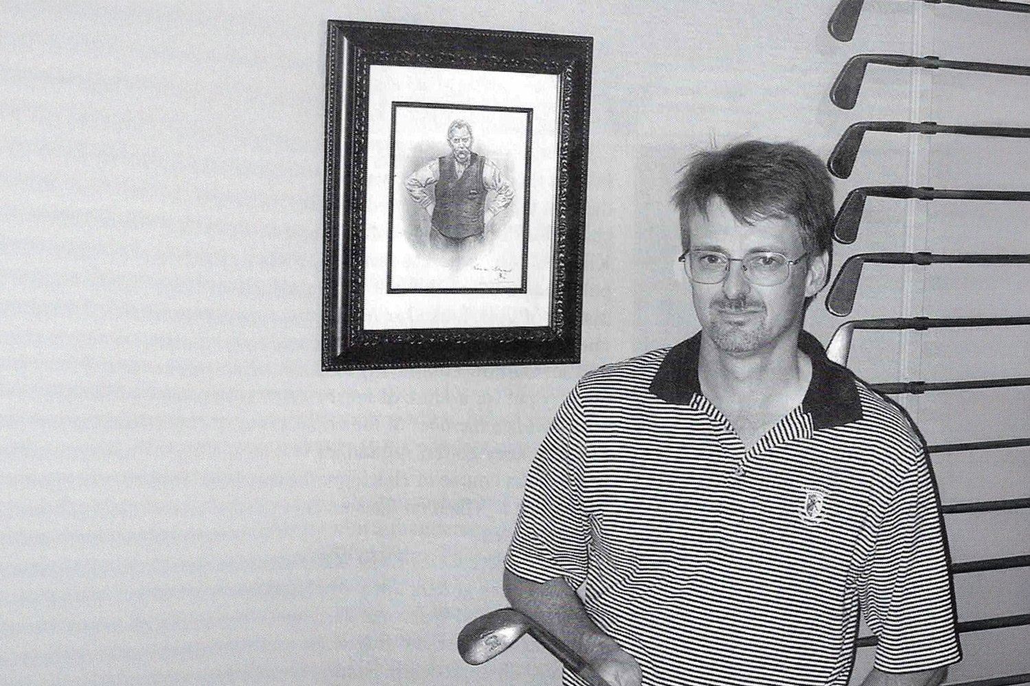 Black-and-white photograph of a man wearing glasses and a striped polo shirt, standing indoors and holding a hickory golf club. Behind him, a framed portrait hangs on the wall, and a vertical display of vintage golf clubs is visible to his side, suggesting a private collection or study space.