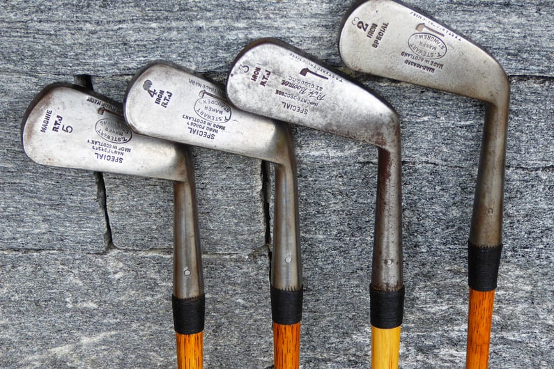 Group of vintage Tom Stewart hickory golf irons displayed side by side, showing different niblick and mashie club heads with hand-forged markings and wooden shafts.