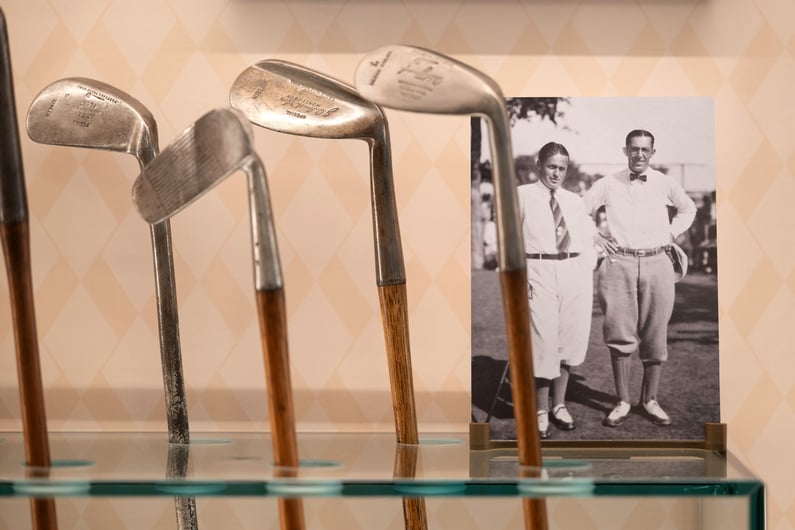 Vintage hickory golf clubs displayed on a glass shelf, with a historic black-and-white photograph of early 20th-century golfers in the background, evoking the material history and tradition of the game.