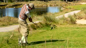 Paolo Quirici hitting a golf shot with a hickory club, dressed in vintage knickerbockers, cap, and patterned vest, on the fairway near a lake at Golf Club Föhr during the German Open Hickory Championship 2025.