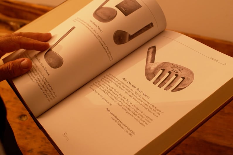Hands holding an open book showing detailed descriptions and images of early golf clubs, including the Higgs Bulmer “Rake” niblick.