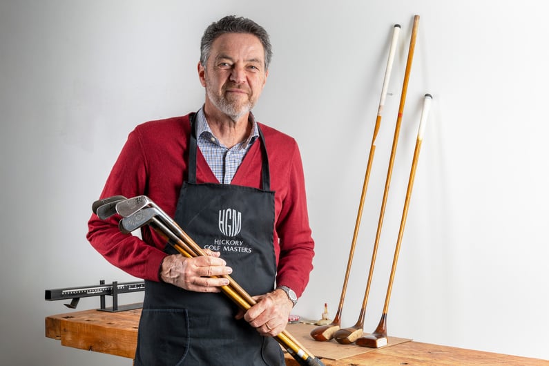 Craftsman holding restored hickory golf clubs in a workshop setting, wearing a Hickory Golf Masters apron, with vintage wooden clubs displayed on a workbench behind him.