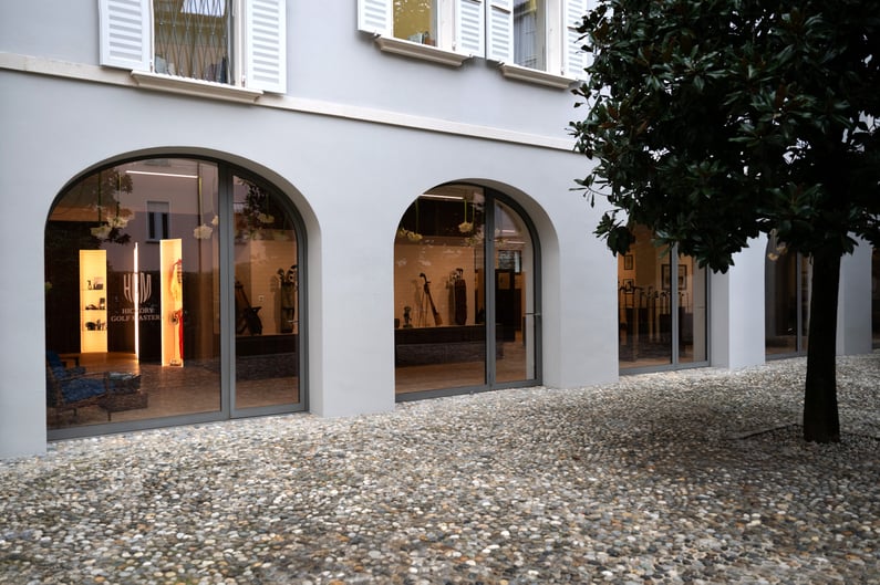 Exterior view of an exhibition space with large arched glass windows, revealing a curated display of historic hickory golf clubs and equipment inside, set within an elegant and refined architectural setting