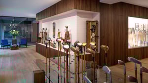 Interior of a golf museum featuring a curated display of vintage wooden golf clubs on a glass stand, with leather golf bags and historic equipment exhibited against warm wood-paneled walls.