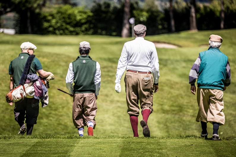 Four golfers dressed in traditional hickory golf attire walk down a fairway on a sunny day. Seen from behind, they wear flat caps, plus-fours, knee-high socks, and vintage-style clothing, with one player carrying a golf bag.
