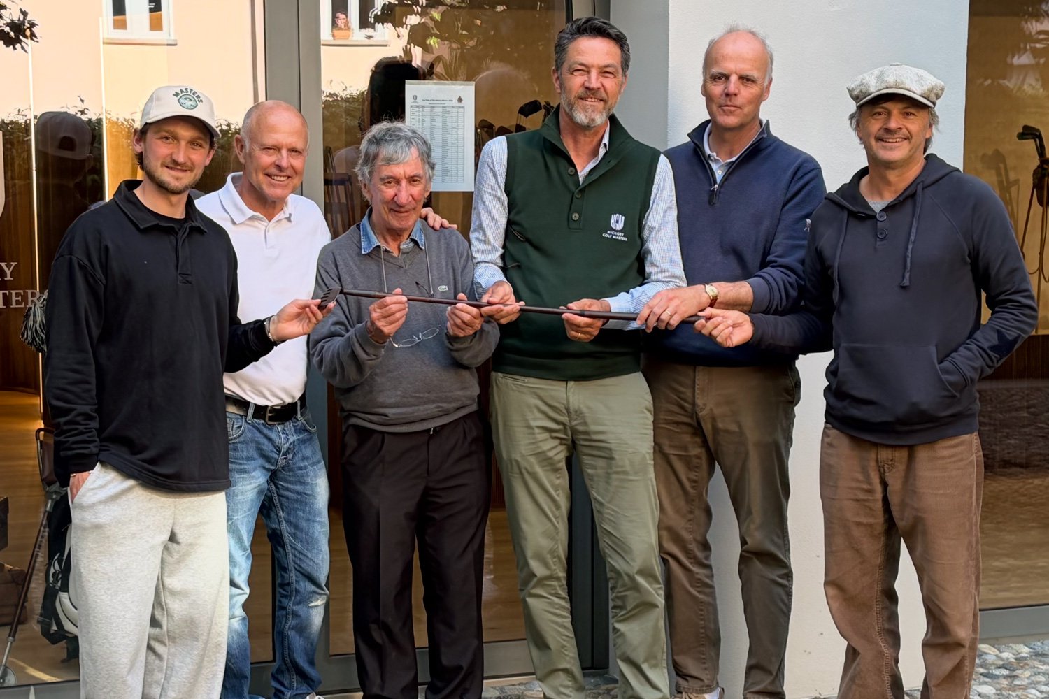 Six men stand together outside a golf clubhouse, holding a vintage hickory golf club and posing for a commemorative photograph. The group includes golfers and enthusiasts of different generations, dressed in a mix of casual and golf attire.