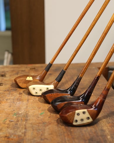 Original wooden-headed hickory golf clubs lined up on a traditional wooden workbench.