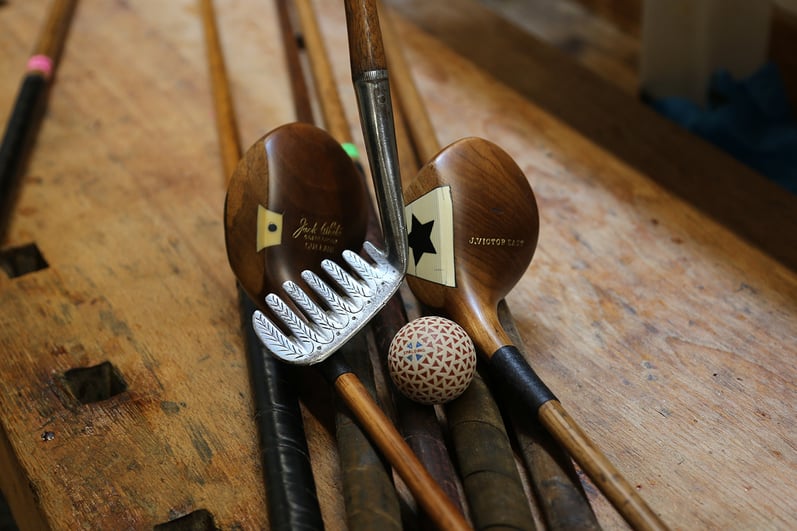 Vintage hickory golf clubs with wooden heads, an antique iron and a period golf ball displayed on a wooden workbench.