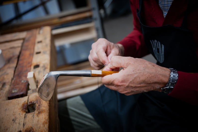 Close-up of a craftsman restoring a hickory golf club, carefully binding the wooden shaft to the metal club head on a workbench in a traditional workshop.