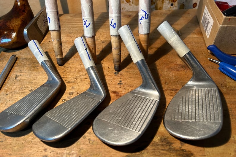 Set of restored hickory iron club heads laid out on a wooden workbench, with shafts marked and prepared for fitting during the restoration process.