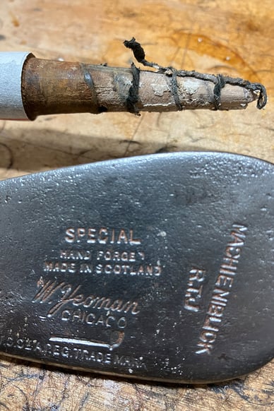 Close-up of a vintage MacGregor hickory iron club head on a wooden workbench, with the shaft wrapped and marked for restoration, showing original stamping and signs of age.