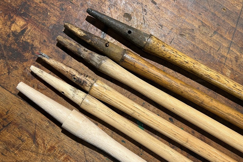 Selection of hickory golf club shafts at different stages of wear and restoration, laid out on a wooden workbench, showing original patina, repairs, and newly shaped wood.