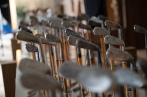 Rows of antique hickory golf clubs standing upright in a workshop, with iron heads aligned and wooden shafts visible, awaiting restoration and finishing.