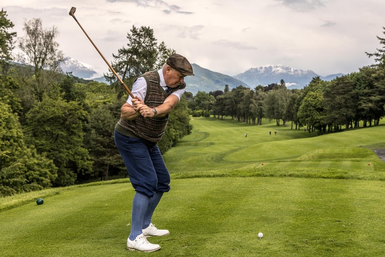 A golfer in traditional hickory-era attire swinging a wooden-shafted club on a mountain golf course.