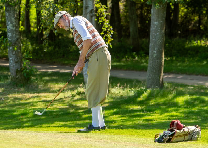 Paolo Quirici wearing traditional hickory golf attire, including cap, vest and knickerbockers, carefully putting on a sunlit green with a vintage golf bag and wooden-shaft clubs resting on the grass nearby.