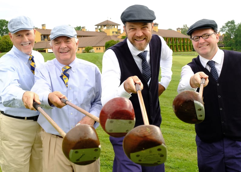 Four golfers wearing vintage hickory golf attire smile and point their wooden-head clubs toward the camera on a golf course, with a clubhouse visible in the background.