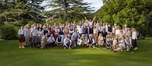 Large group of golfers wearing traditional hickory golf attire posing together on a lush golf course, surrounded by tall trees and greenery on a sunny day, with vintage clubs and period clothing including caps, vests, and knickerbockers