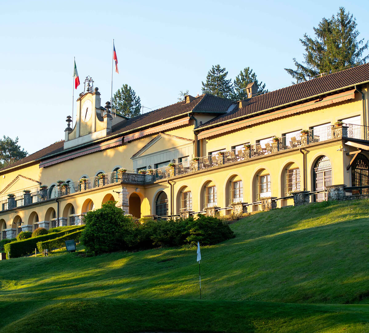 Elegant clubhouse of Golf Club Villa d’Este, nestled in the hills, with Italian and Czech flags and a lush green fairway in the foreground.