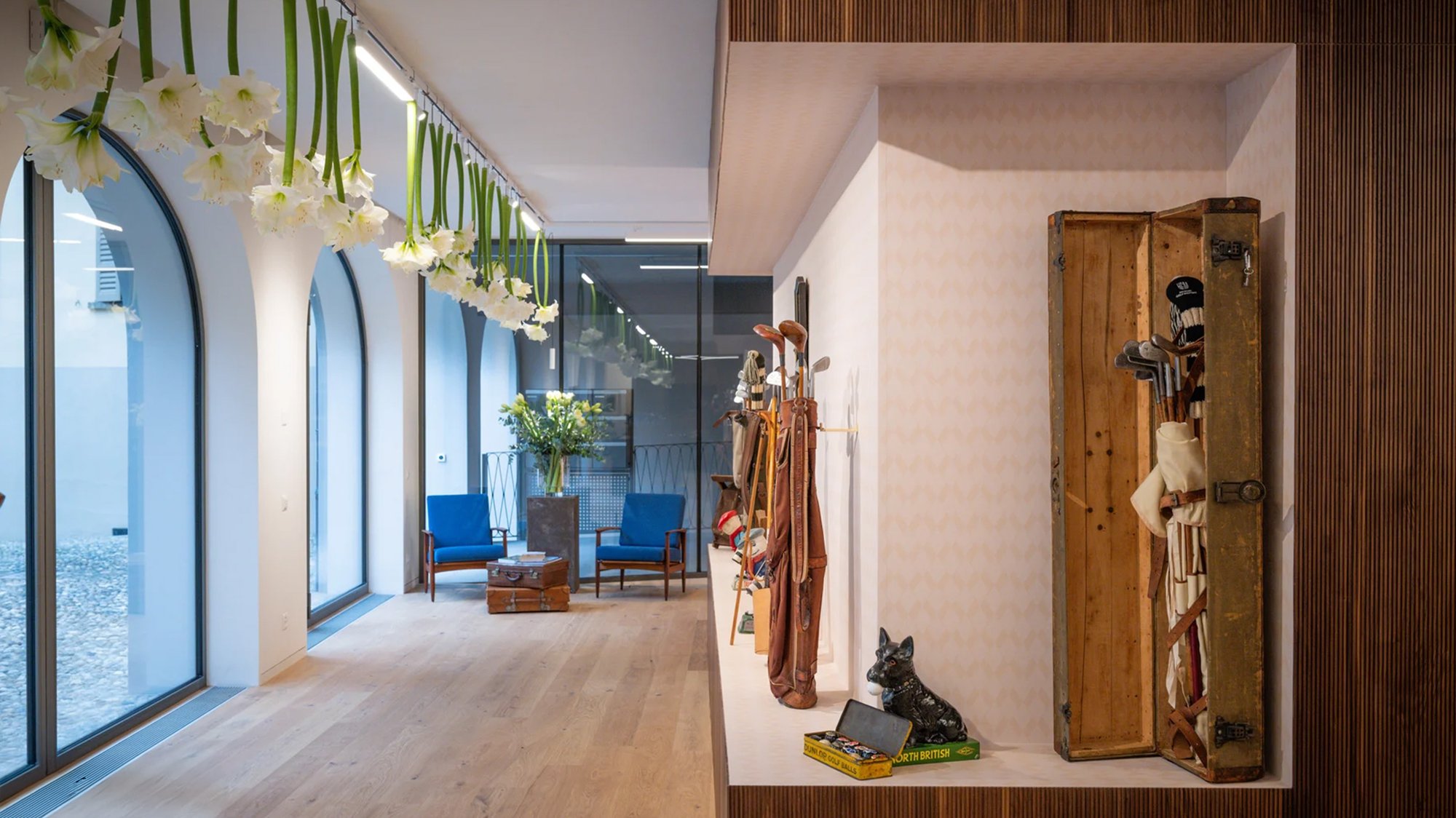Wide interior view of the Hickory Golf Masters Museum, featuring hanging white flowers, large arched windows, blue lounge chairs, and a display of vintage golf bags and wooden-shaft clubs set into a recessed wall. Wide interior view of the Hickory Golf Masters Museum, featuring hanging white flowers, large arched windows, blue lounge chairs, and a display of vintage golf bags and wooden-shaft clubs set into a recessed wall.